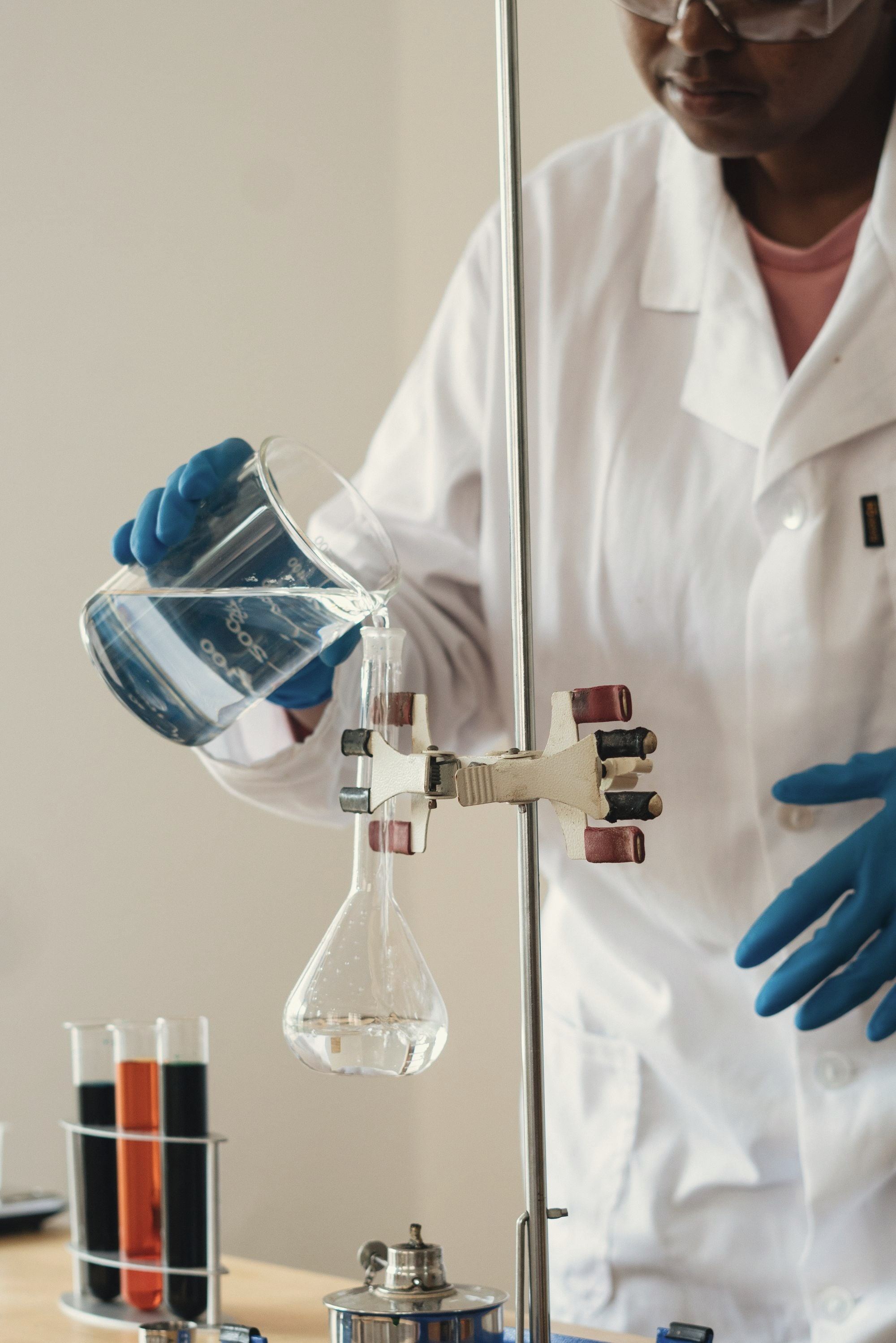 Gloved hands pouring liquid from a beaker into a flask on a lab stand